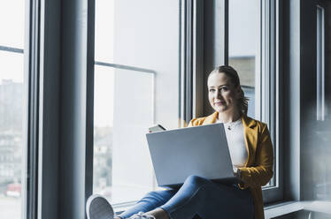 Smiling businesswoman with laptop sitting near window at office - UUF28457