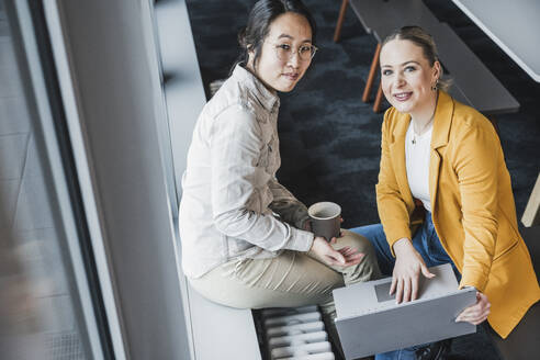 Lächelnde Geschäftsfrauen sitzen am Fenster im Büro - UUF28460