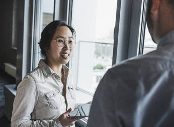 Businesswoman wearing eyeglasses discussing with colleague near window at office - UUF28461