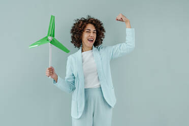 Happy woman holding wind turbine model flexing muscles against blue background - YTF00588