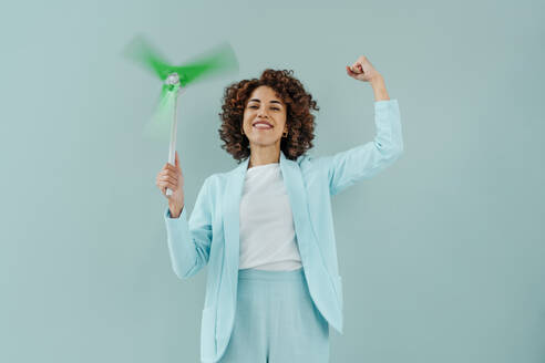 Happy woman flexing muscles and holding wind turbine spinning against blue background - YTF00589