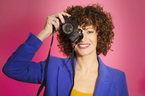 Lächelnde Frau beim Fotografieren mit Kamera vor magentafarbenem Hintergrund - JSMF02730