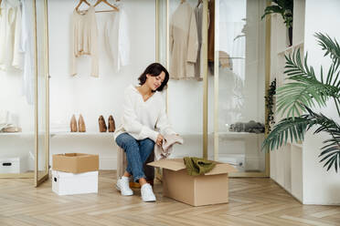 Young woman putting clothes in box sitting in front of closet at home - VPIF07890