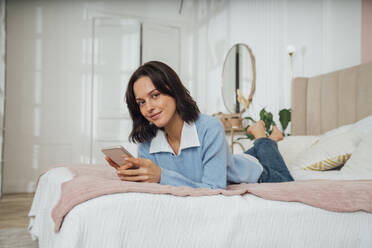 Lächelnde junge Frau auf dem Bett liegend mit Smartphone zu Hause - VPIF07918