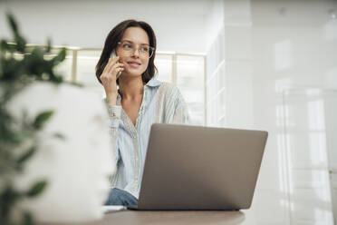 Smiling freelancer talking on smart phone and sitting with laptop at desk - VPIF07935