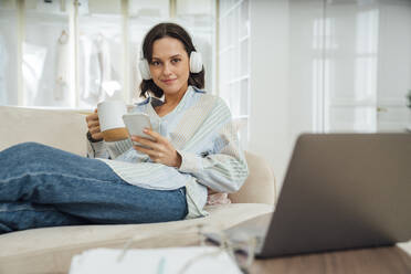 Smiling freelancer wearing wireless headphones sitting with smart phone and coffee cup on sofa at home - VPIF07938