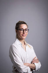 Smiling businesswoman wearing eyeglasses with arms crossed standing against gray background - MIKF00125