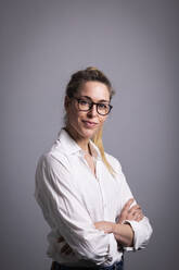 Smiling businesswoman wearing eyeglasses standing with arms crossed against gray background - MIKF00126