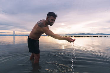 Glücklicher Mann spielt mit Wasser und genießt den Sonnenuntergang am Strand - PNAF05019
