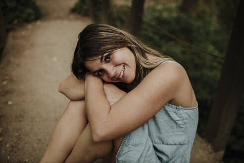 Glückliche junge Frau sitzt auf einem Fußweg im Wald - GMLF01315