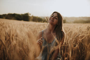 Happy young woman with dry plant sitting in field - GMLF01321