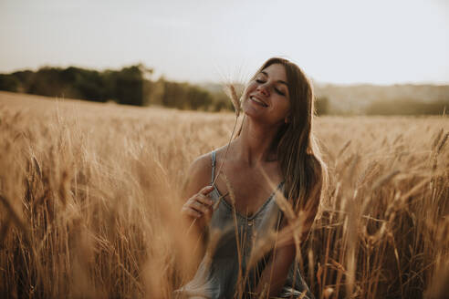 Glückliche junge Frau mit trockener Pflanze im Feld sitzend - GMLF01321