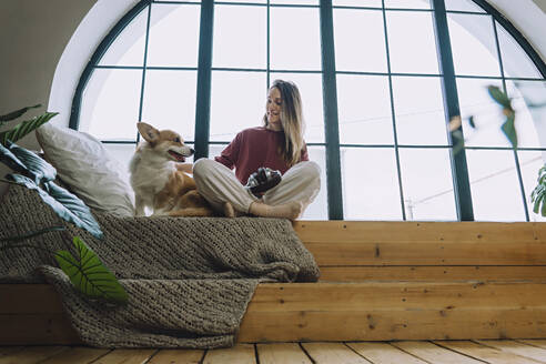 Smiling woman sitting cross-legged with dog on window sill at home - IEF00318