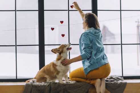 Frau klebt Herzaufkleber auf Fenster und sitzt bei ihrem Hund zu Hause - IEF00323