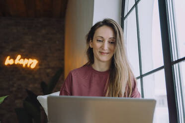 Smiling woman watching laptop near window at home - IEF00337