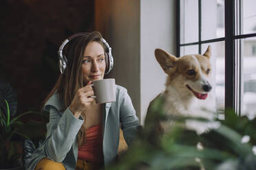 Frau mit Kopfhörern trinkt Kaffee mit Hund zu Hause - IEF00344