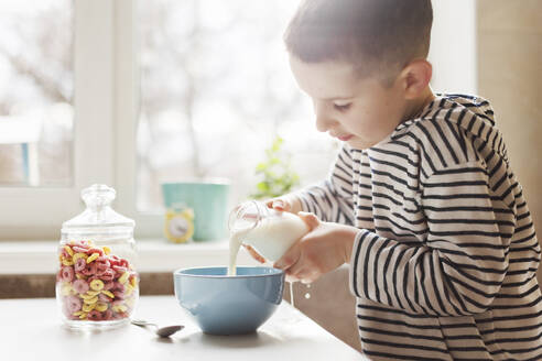 Boy pouring milk in bowl on table at home - ONAF00430