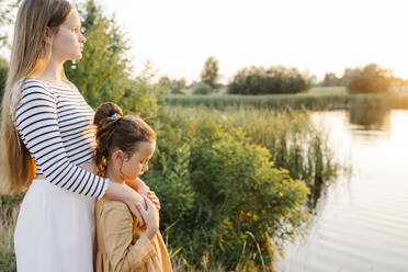 Nachdenkliches Mädchen mit Schwester am See bei Sonnenuntergang stehend - SSYF00146