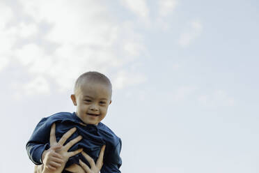 Niedlicher blonder Junge mit Down-Syndrom auf Mamas Armen, blauer Himmel Hintergrund - CAVF96595