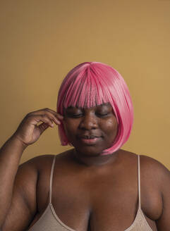 Plus size woman wearing pink bob wig posing with eyes closed against beige background - VRAF00080