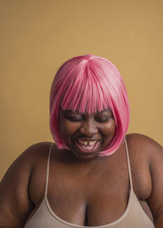 Cheerful plus size woman wearing pink bob wig against beige background - VRAF00081