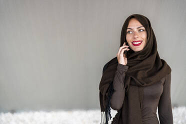 Frau mit Hidschab, die vor einer Wand mit ihrem Smartphone spricht - DLTSF03577