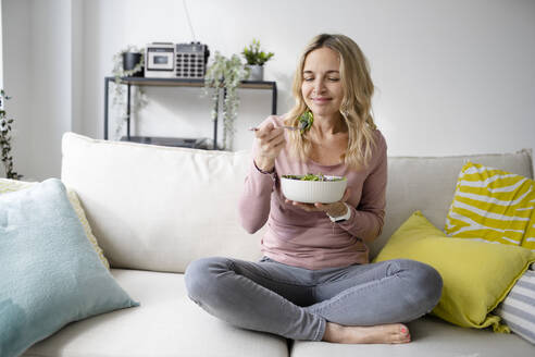 Smiling woman with bowl of salad sitting on sofa - HMEF01520
