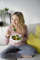Thoughtful woman sitting with bowl of salad on sofa - HMEF01521