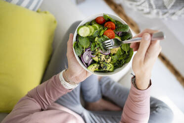 Hands of woman with bowl of salad sitting on sofa at home - HMEF01523