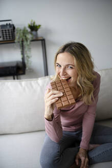 Glückliche Frau isst Schokolade auf dem Sofa zu Hause - HMEF01524
