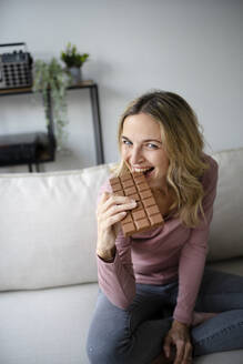 Happy woman eating chocolate on sofa at home - HMEF01524