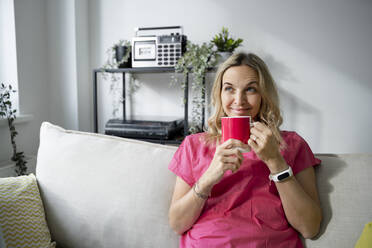 Smiling mature woman with coffee cup sitting on sofa at home - HMEF01525