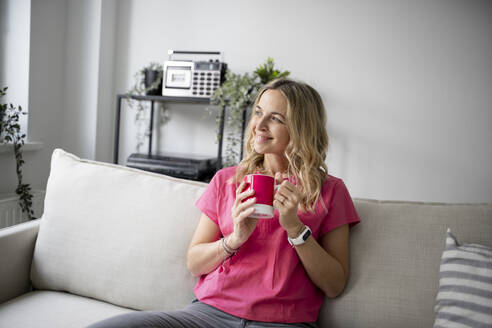 Lächelnde reife Frau mit Kaffeetasse auf dem Sofa zu Hause sitzend - HMEF01527