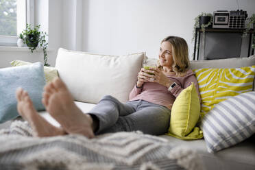 Lächelnde Frau sitzt mit einem Glas Smoothie auf dem Sofa zu Hause - HMEF01529