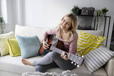 Glückliche reife Frau singt und spielt Gitarre auf dem Sofa zu Hause - HMEF01530
