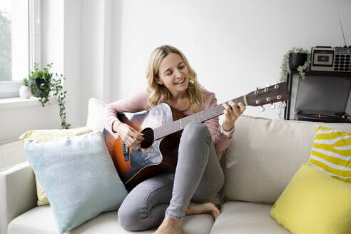 Lächelnde Frau spielt Gitarre auf dem Sofa zu Hause - HMEF01532