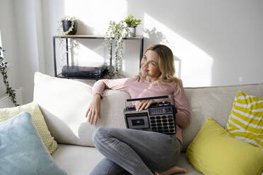 Smiling mature woman sitting with radio on sofa at home - HMEF01538