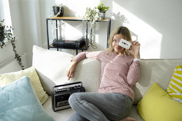 Smiling woman with cassette and radio sitting on sofa at home - HMEF01539