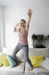 Happy woman wearing wireless headphones listening to music and dancing on sofa at home - HMEF01542
