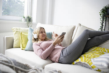 Mature woman wearing wireless headphones lying on sofa at home - HMEF01549