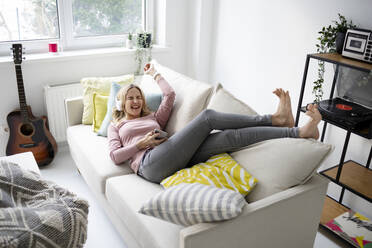 Cheerful woman wearing wireless headphones lying on sofa at home - HMEF01551