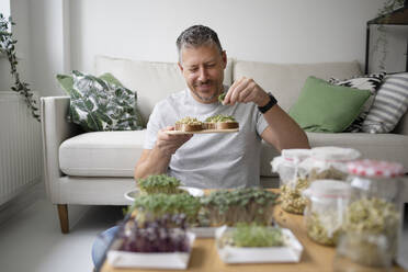 Smiling man eating sprouts sitting in front of sofa at home - HMEF01553