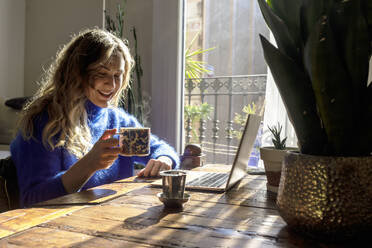 Frau hält eine Tasse Tee und arbeitet zu Hause am Laptop - RFTF00378
