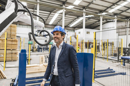 Happy mature engineer looking at robotic arm in factory - JCCMF09676