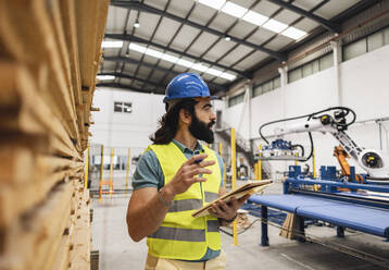 Ingenieur mit Notizblock vor einem Stapel von Brettern in einer Fabrik - JCCMF09710