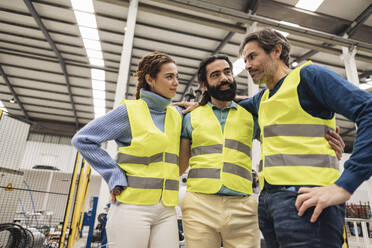 Glückliche Ingenieure in reflektierender Kleidung, die mit dem Arm in einer Roboterfabrik stehen - JCCMF09751