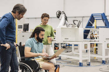 Engineer sitting in wheelchair discussing with colleagues on laptop in factory - JCCMF09788