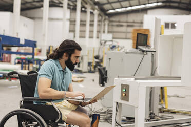 Ingenieur im Rollstuhl sitzend, der in einer Fabrik eine Maschine über einen Laptop untersucht - JCCMF09789