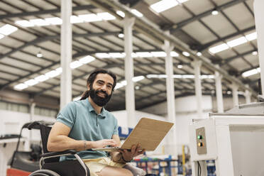 Happy engineer sitting in wheelchair working on laptop in robotics factory - JCCMF09792