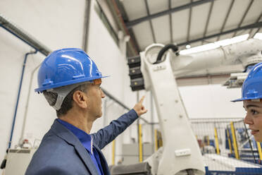 Reifer Geschäftsmann, der auf einen Roboterarm in einer Fabrik zeigt - JCCMF09806